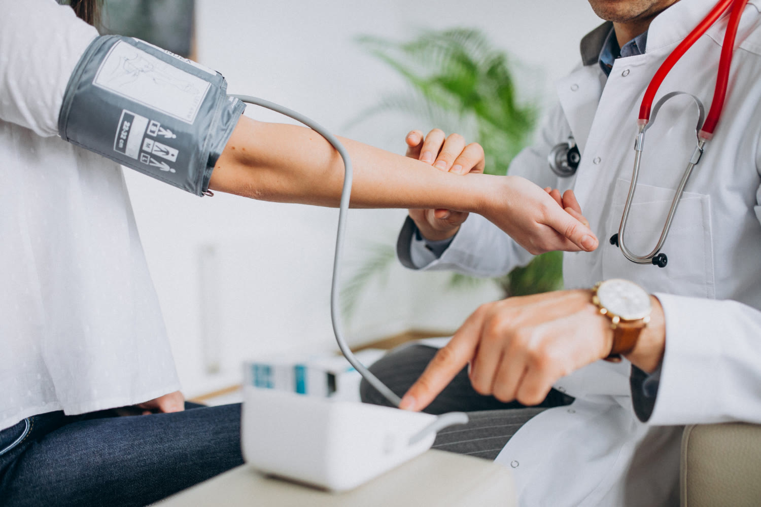 A doctor in a white coat taking a patient's blood pressure, holding their wrist to time the reading while the monitor sits on the table between them.