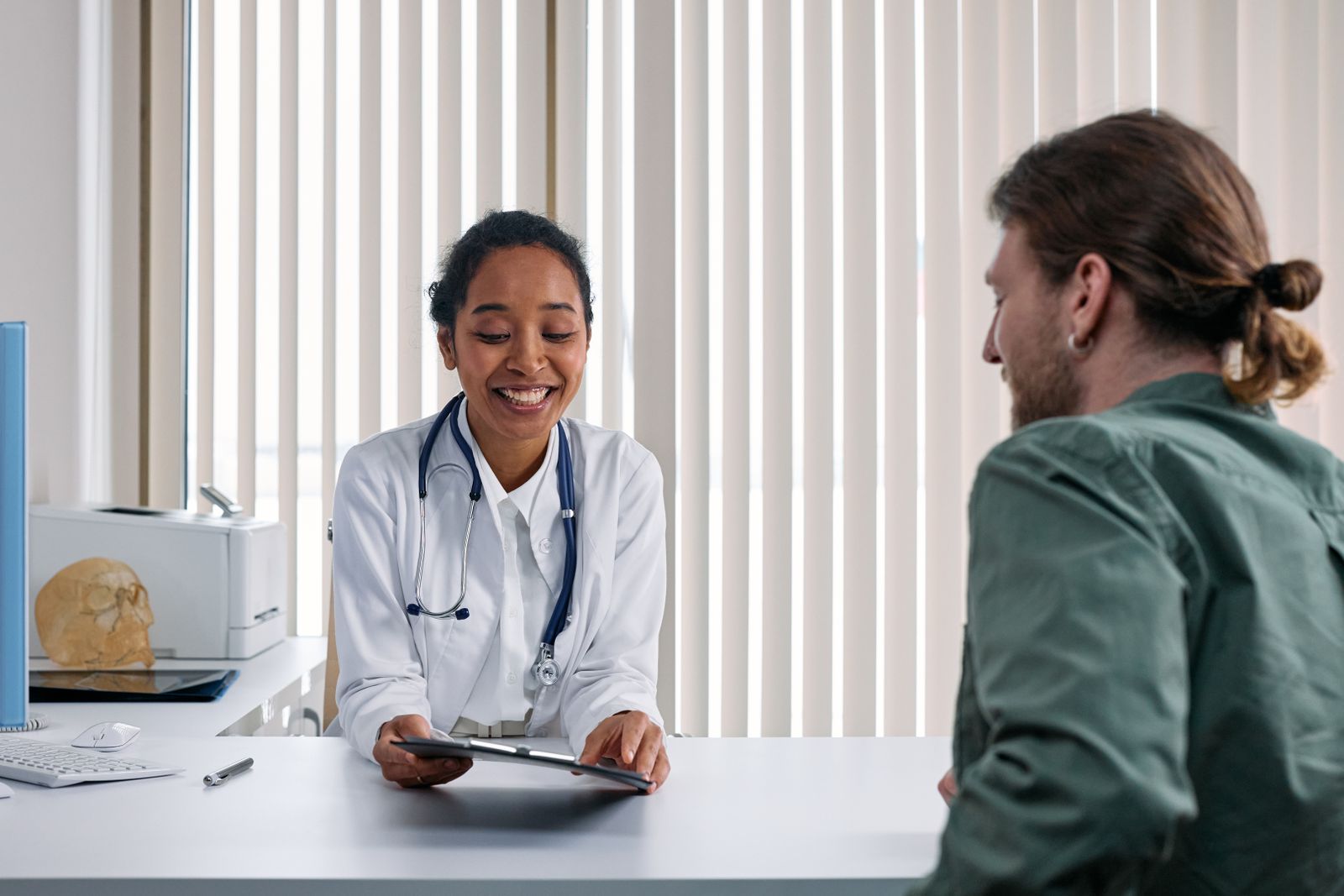 A smiling doctor going through results on a tablet with a patient across a consulting-room desk.